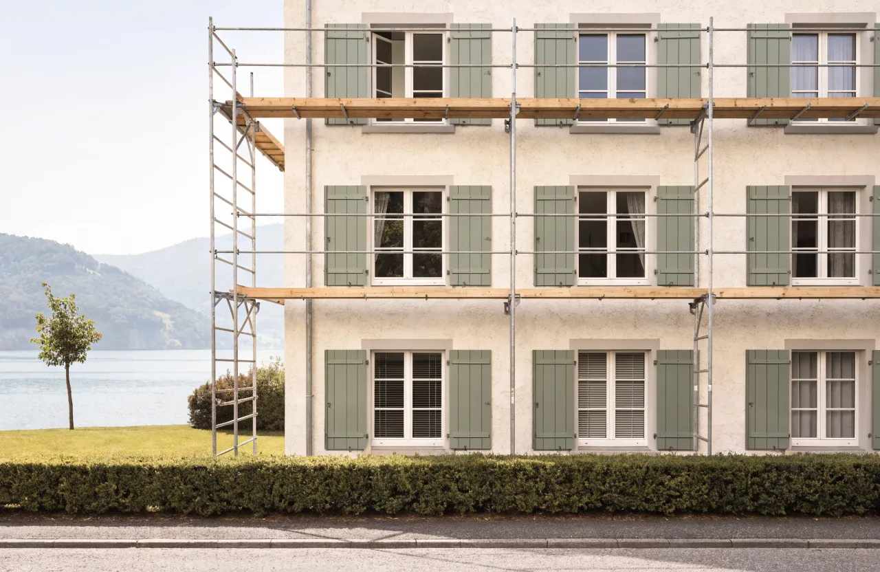 Dieselbe Fassade nach dem Anstrich: glatter Putz in Warmweiss, Fensterläden in gedämpftem Grün, Gerüst noch stehend.