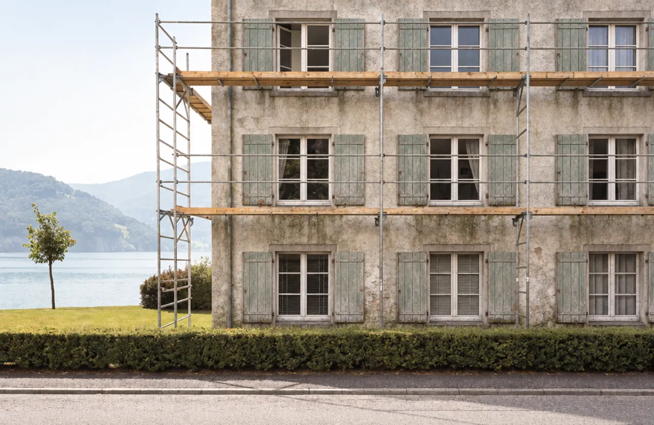 Verwitterte Fassade eines Mehrfamilienhauses mit Gerüst: ausgewaschener Putz, Wasserspuren, abblätternde Fensterläden.